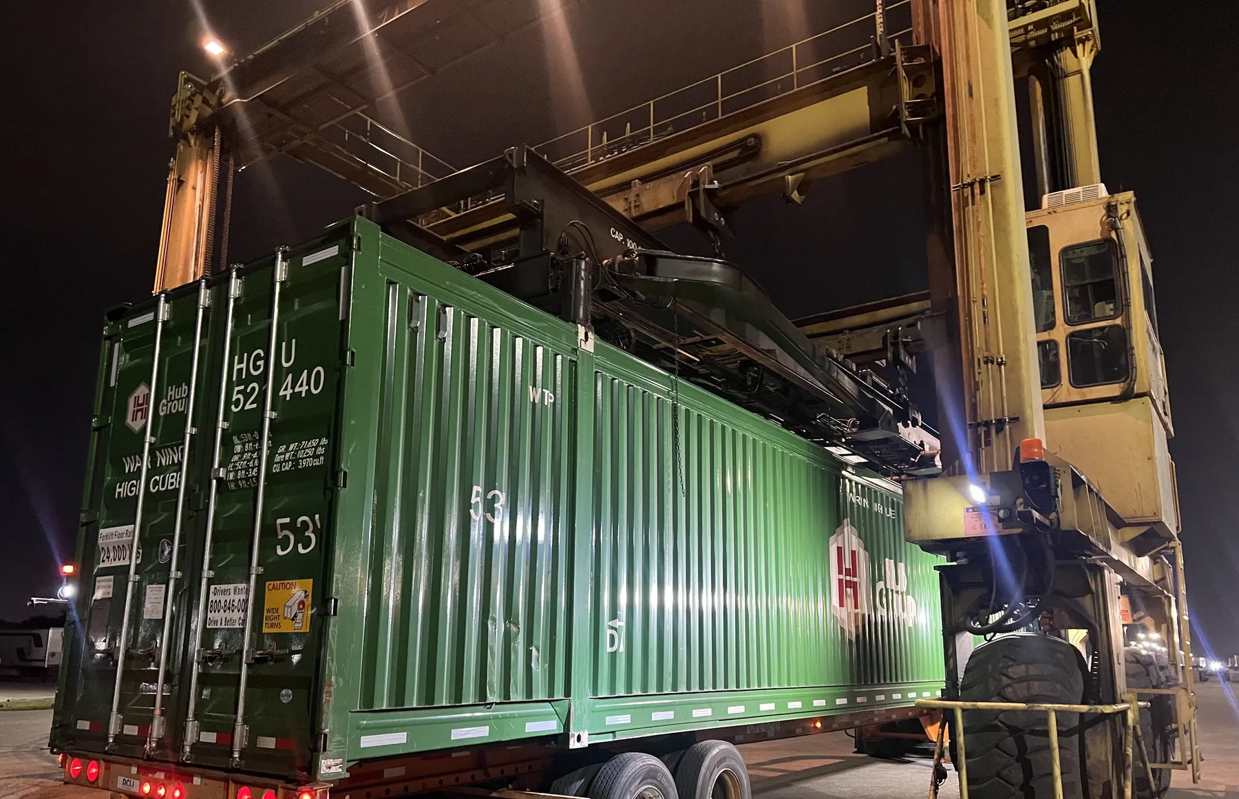 Straddle carrier lifting a green 53-foot container onto a chassis at night